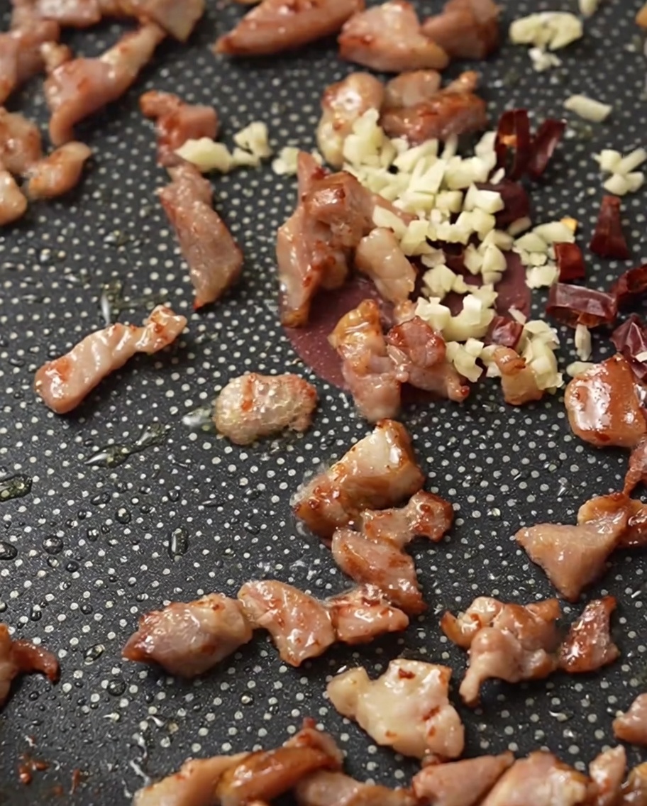Tocino en cubos, ajo picado y chiles rojos secos sofriéndose en una sartén.