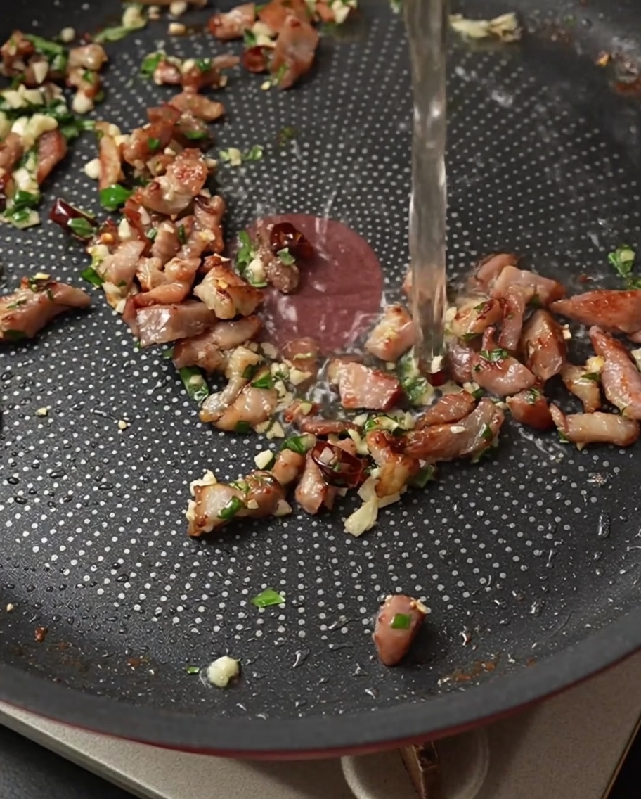 Vino blanco vertiéndose en una sartén con tocino chisporroteante, ajo, chile y albahaca.