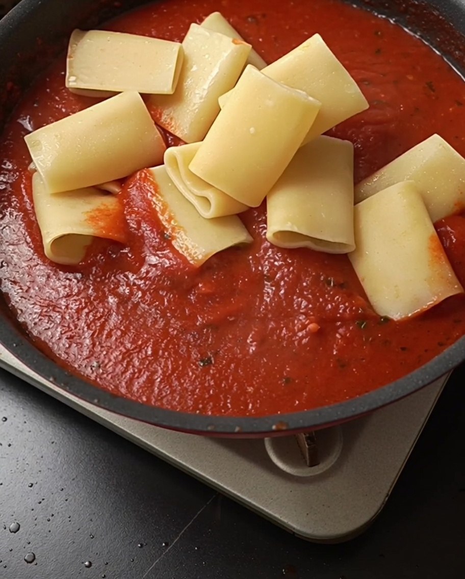 Tubos de pasta cocida añadiéndose a una sartén llena de vibrante salsa de tomate.