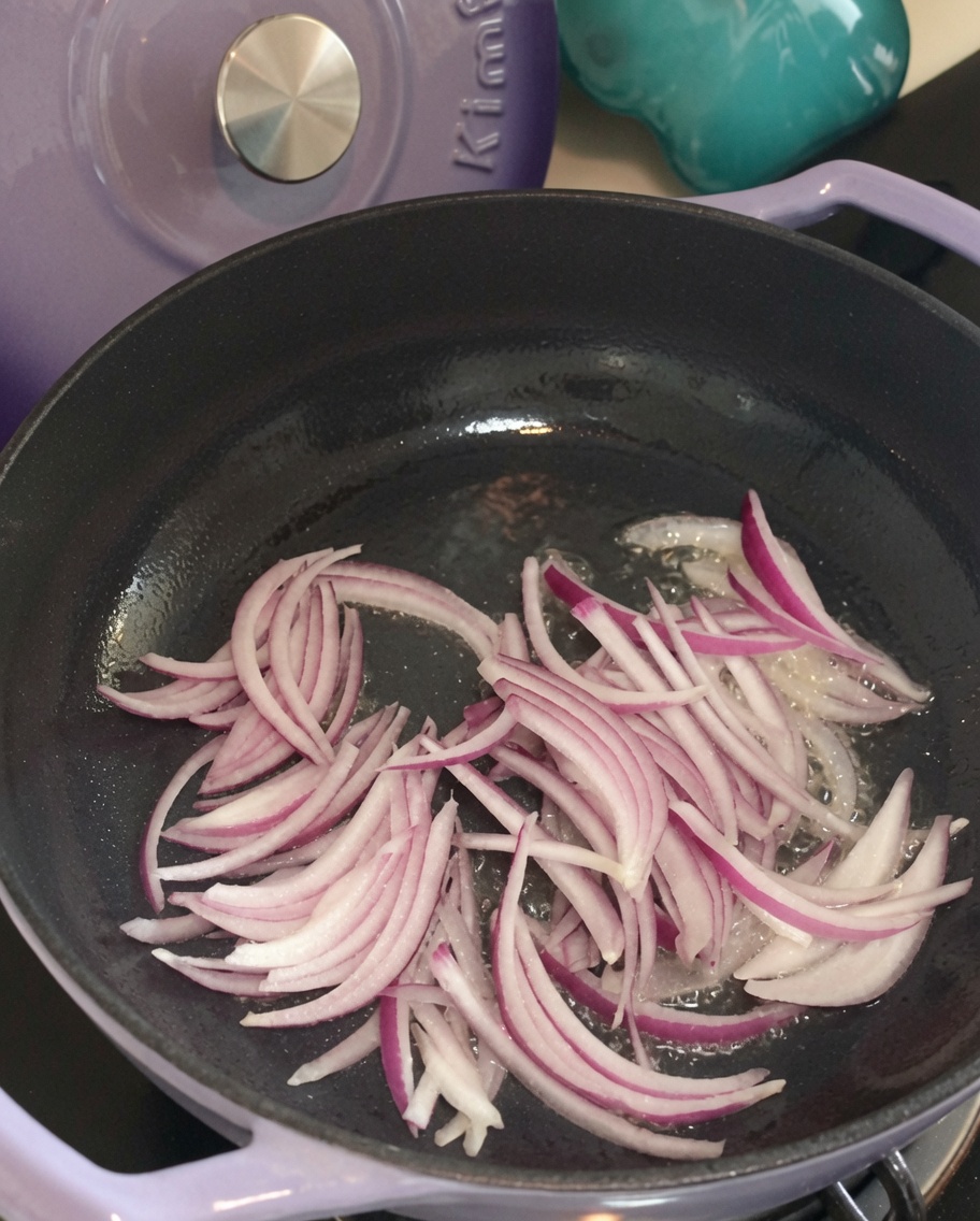 Cebolla roja en rodajas friéndose en una sartén con aceite caliente, empezando a ablandarse.