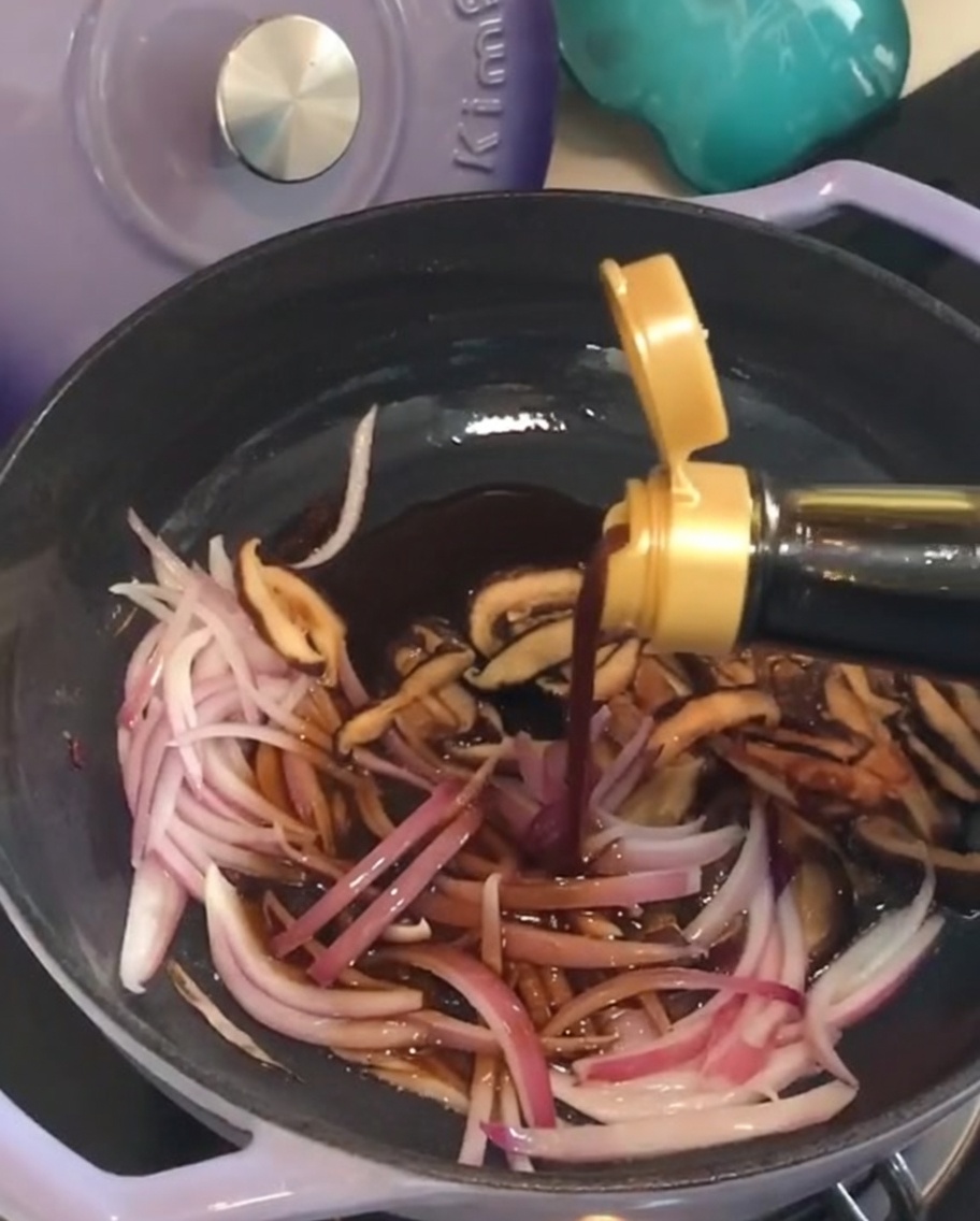 Vertiendo salsa oscura de una botella en una sartén con cebolla roja salteada y setas shiitake.