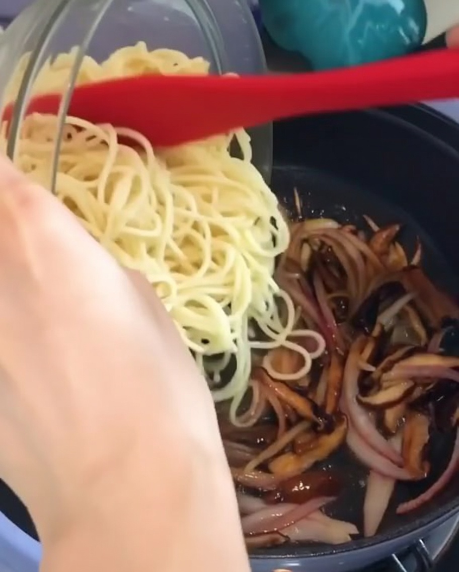 Añadiendo espaguetis cocidos desde un bol de cristal a una sartén con cebollas y setas ya cocinadas.