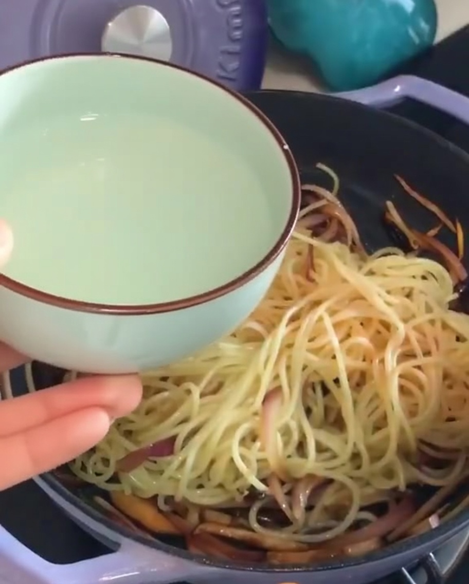 Vertiendo un pequeño cuenco de agua de pasta almidonada en una sartén con espaguetis y verduras.