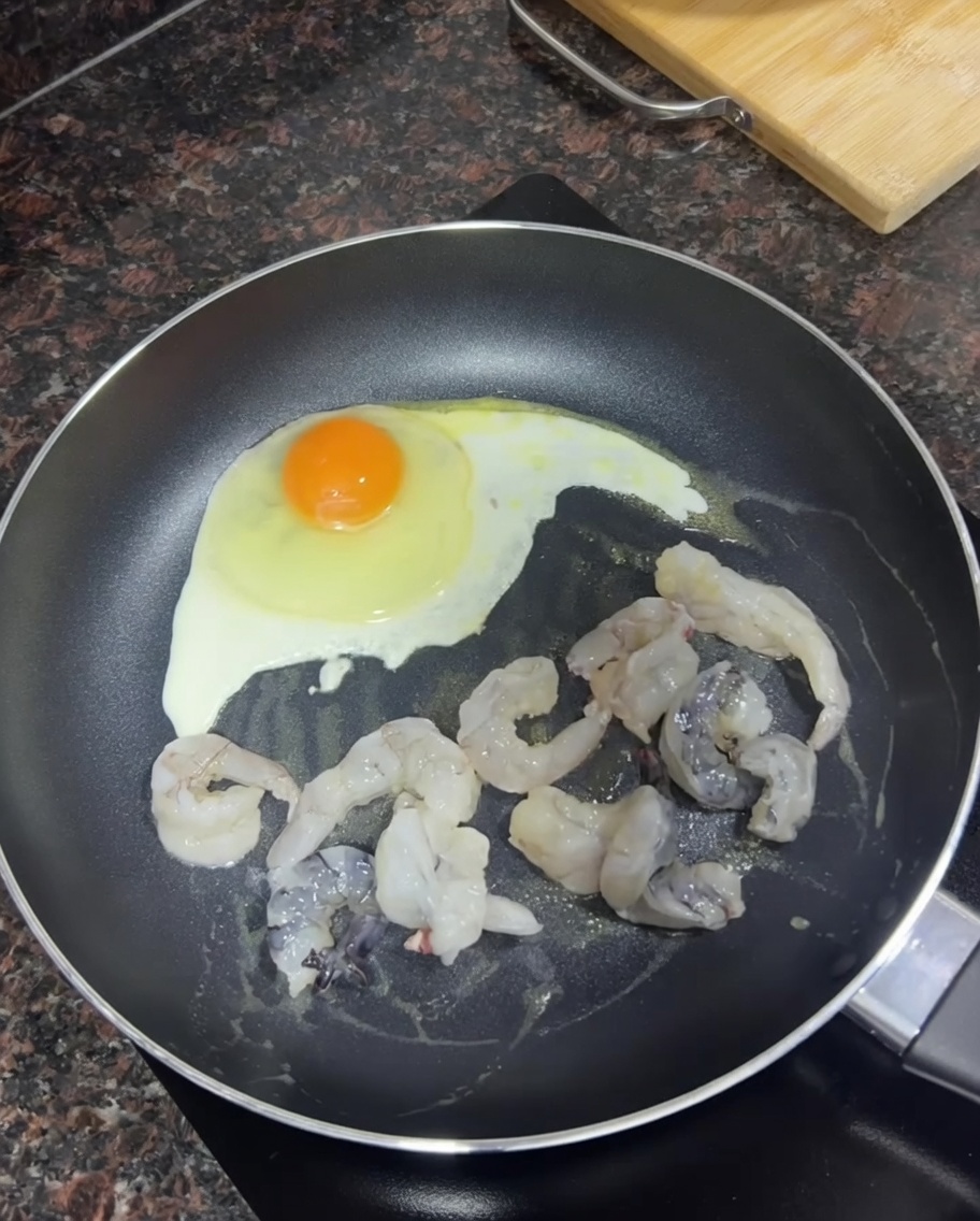 Camarones crudos dorándose en una sartén junto a un huevo friéndose.
