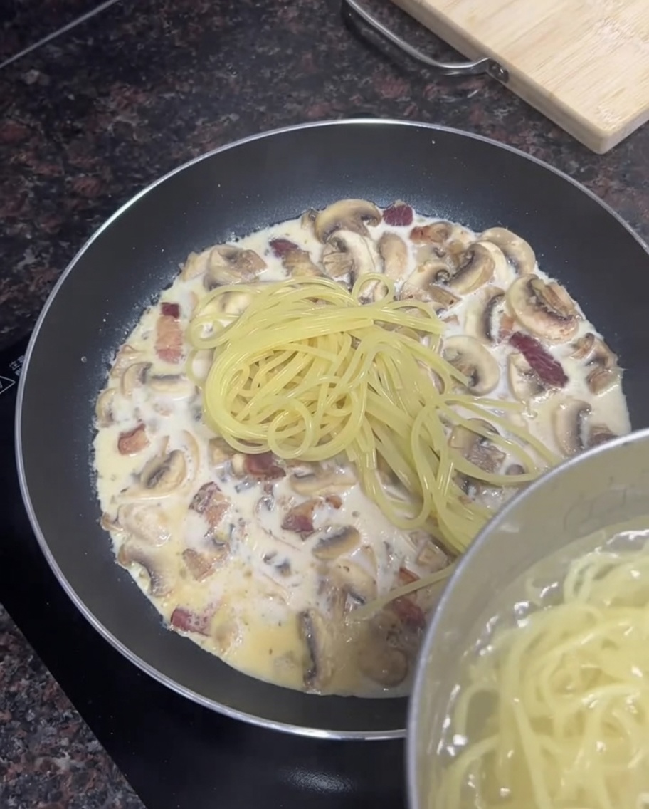 Fideos de espagueti cocidos siendo agregados de una olla a una sartén llena de salsa cremosa de tocino y champiñones.