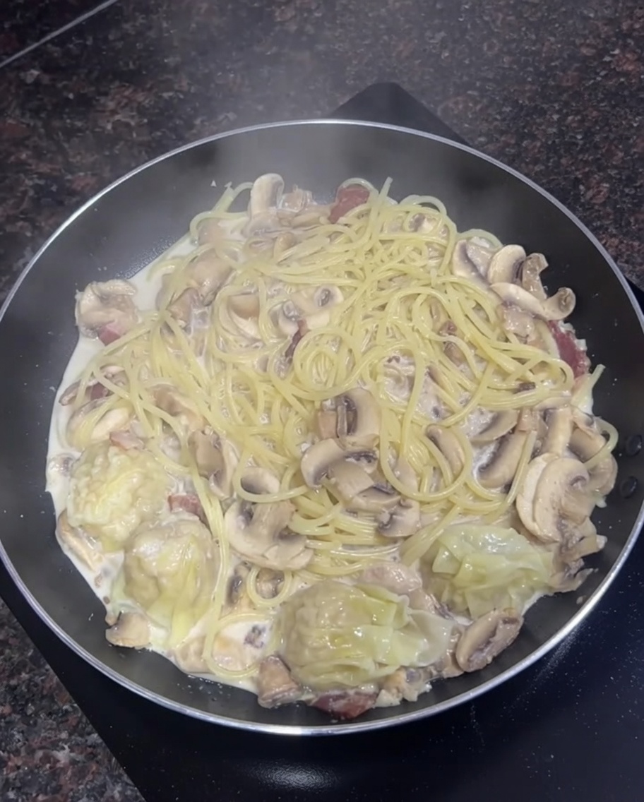 Sartén llena de espaguetis cremosos, champiñones rebanados y unos pocos wontons cocidos hirviendo juntos.