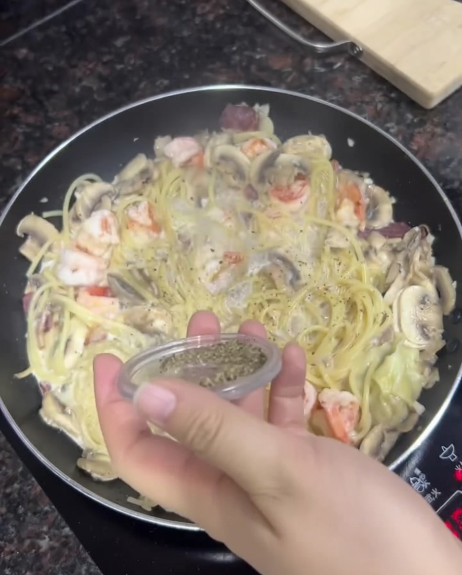Una mano espolvoreando pimienta negra fresca de un recipiente sobre una sartén de pasta cremosa, camarones y champiñones.