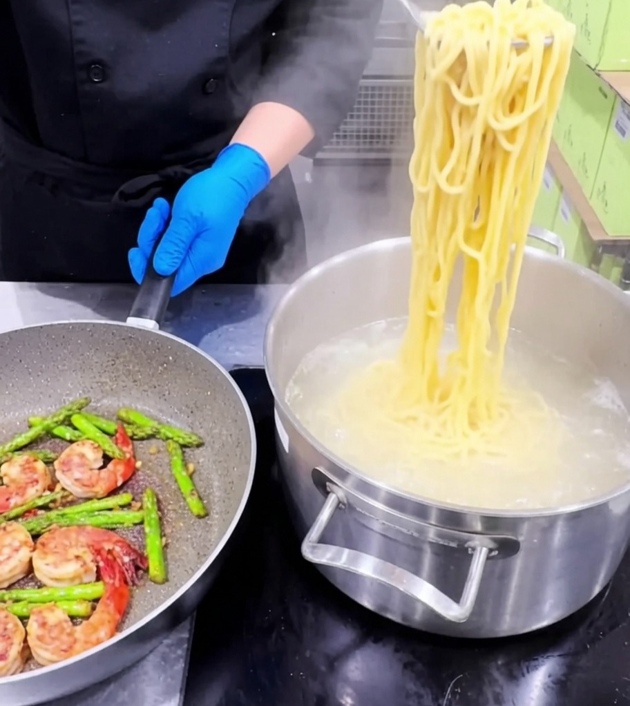 Pasta siendo añadida a una sartén que contiene los camarones, espárragos y ajo cocinados.