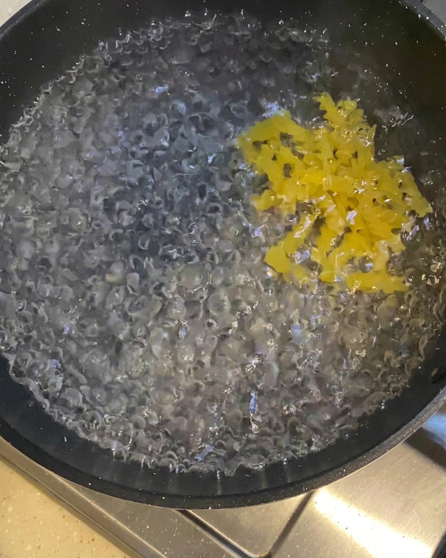 Pasta corta amarilla siendo agregada a una olla oscura llena de agua hirviendo vigorosamente.