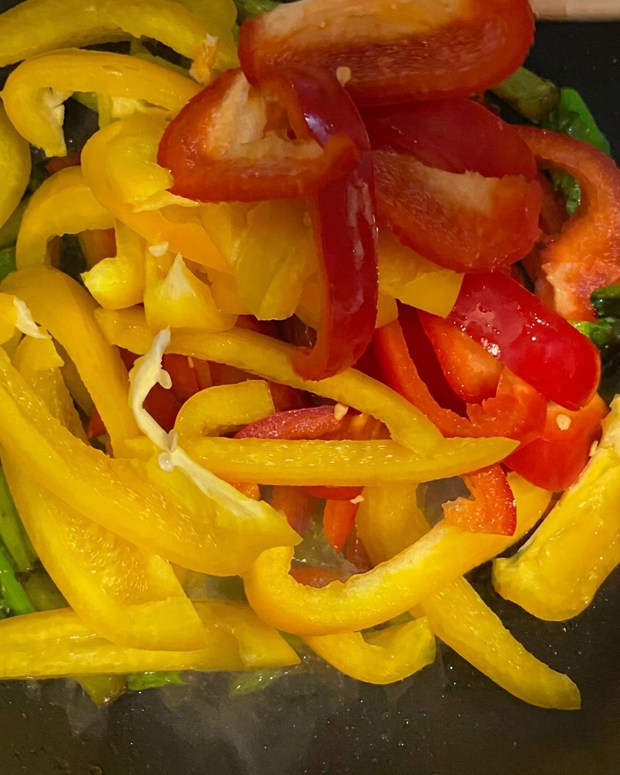 Tiras de pimiento rojo y amarillo vibrante siendo agregadas a una sartén con espárragos verdes.