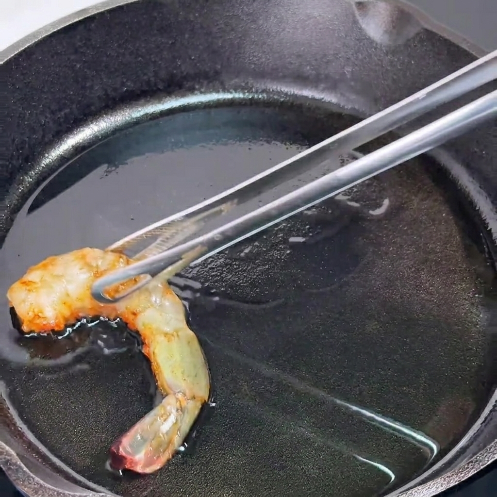Pinzas de metal sosteniendo una gamba parcialmente cocinada sobre una sartén de hierro fundido caliente y aceitada.