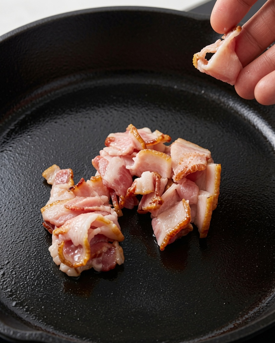 Una mano dejando caer trozos de bacon crudo en rodajas en una sartén de hierro fundido oscura.