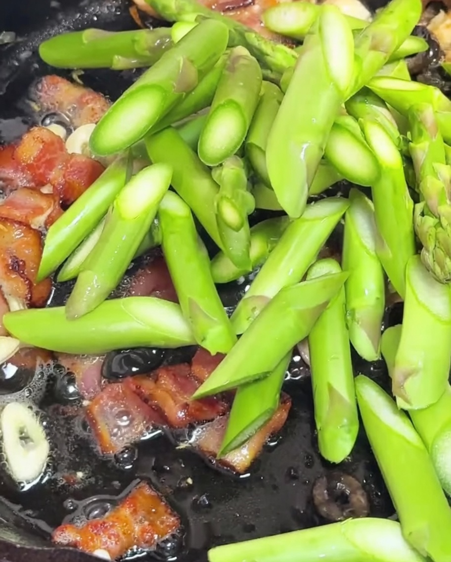 Trozos de espárragos y tomates cherry partidos siendo salteados en una sartén con bacon y ajo.