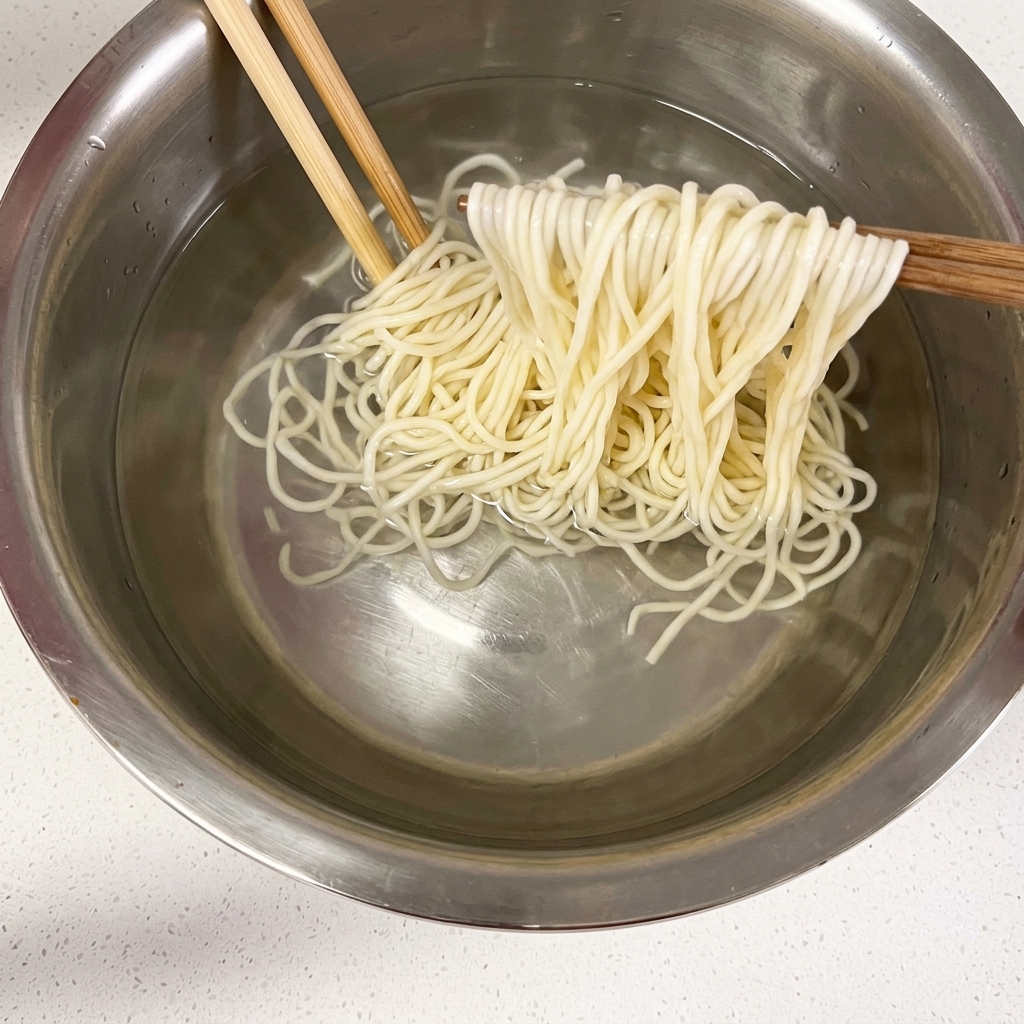 Fideos cocidos sostenidos con palillos sobre un cuenco con agua fría.