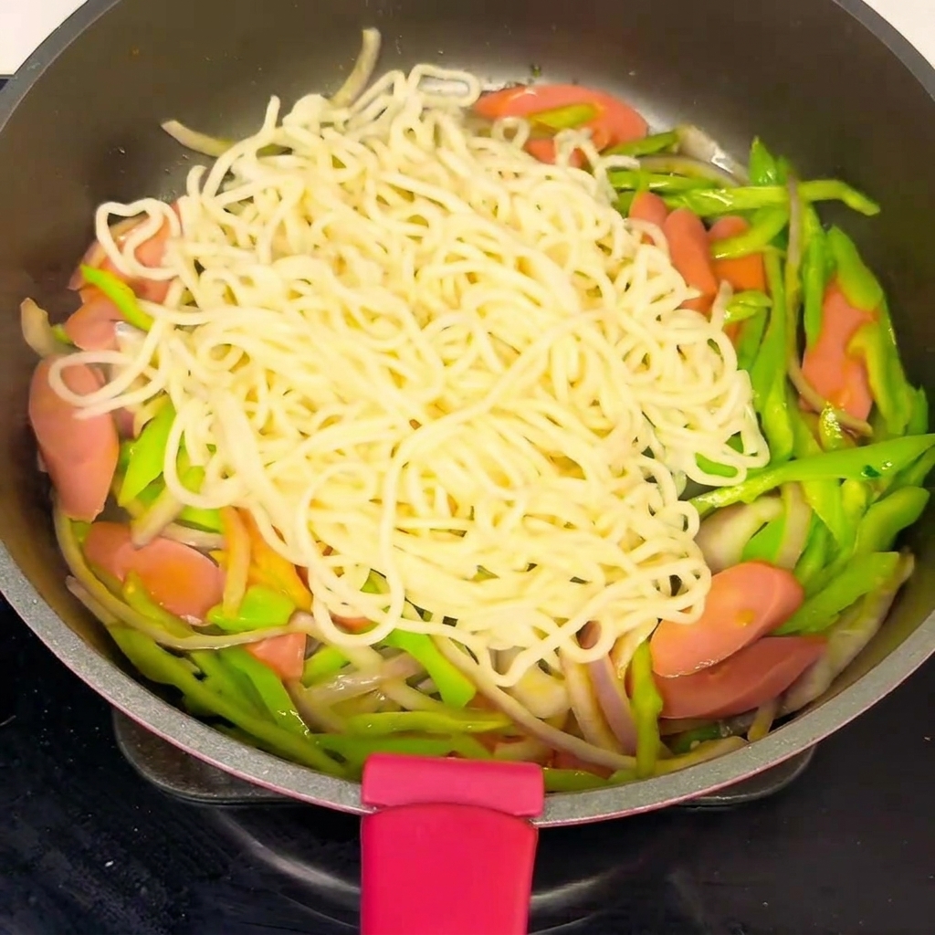 Una sartén llena de fideos y verduras, con la salsa siendo vertida sobre la mezcla.