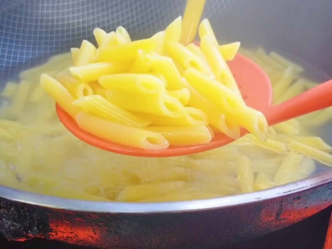 Cuchara ranurada de silicona roja levantando pasta penne cocida del agua hirviendo.