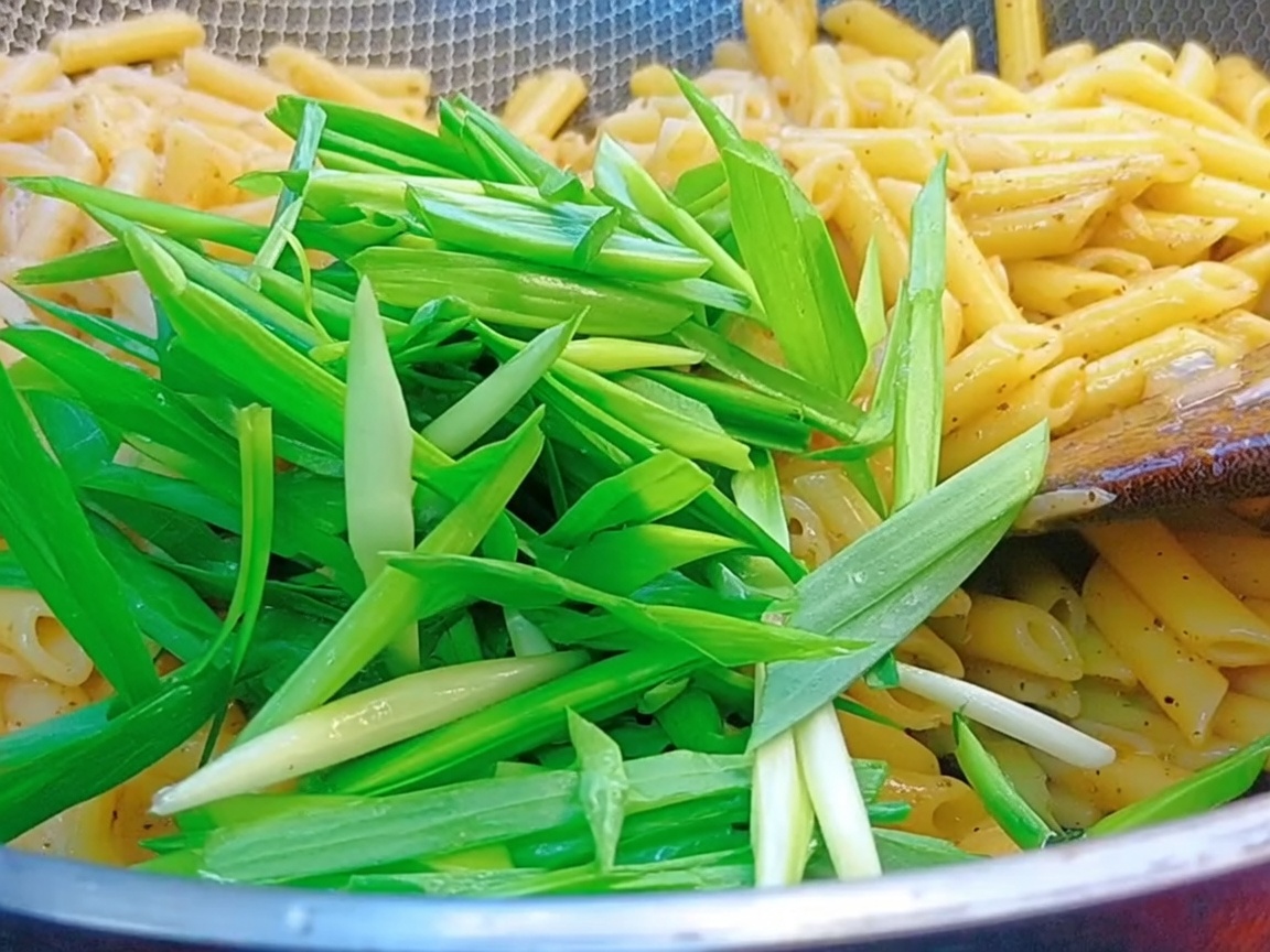 Brotes de ajo verdes frescos siendo echados a un wok lleno de pasta penne y salsa de pimienta negra.