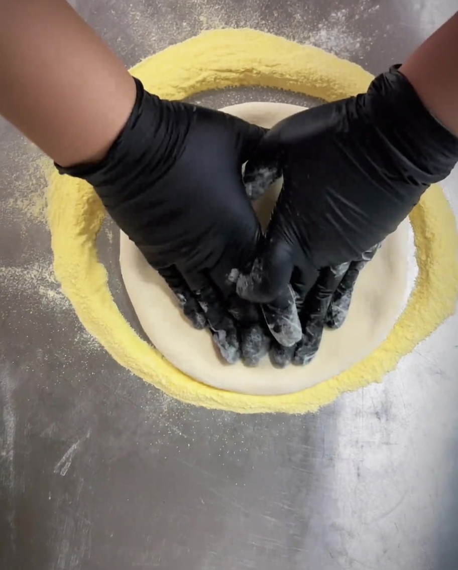Manos con guantes negros presionando el centro de la masa para aplanarla uniformemente.