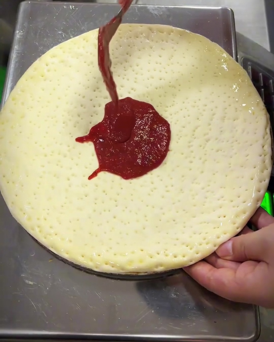 Salsa de tomate siendo extendida sobre la masa con el dorso de una cuchara de metal.