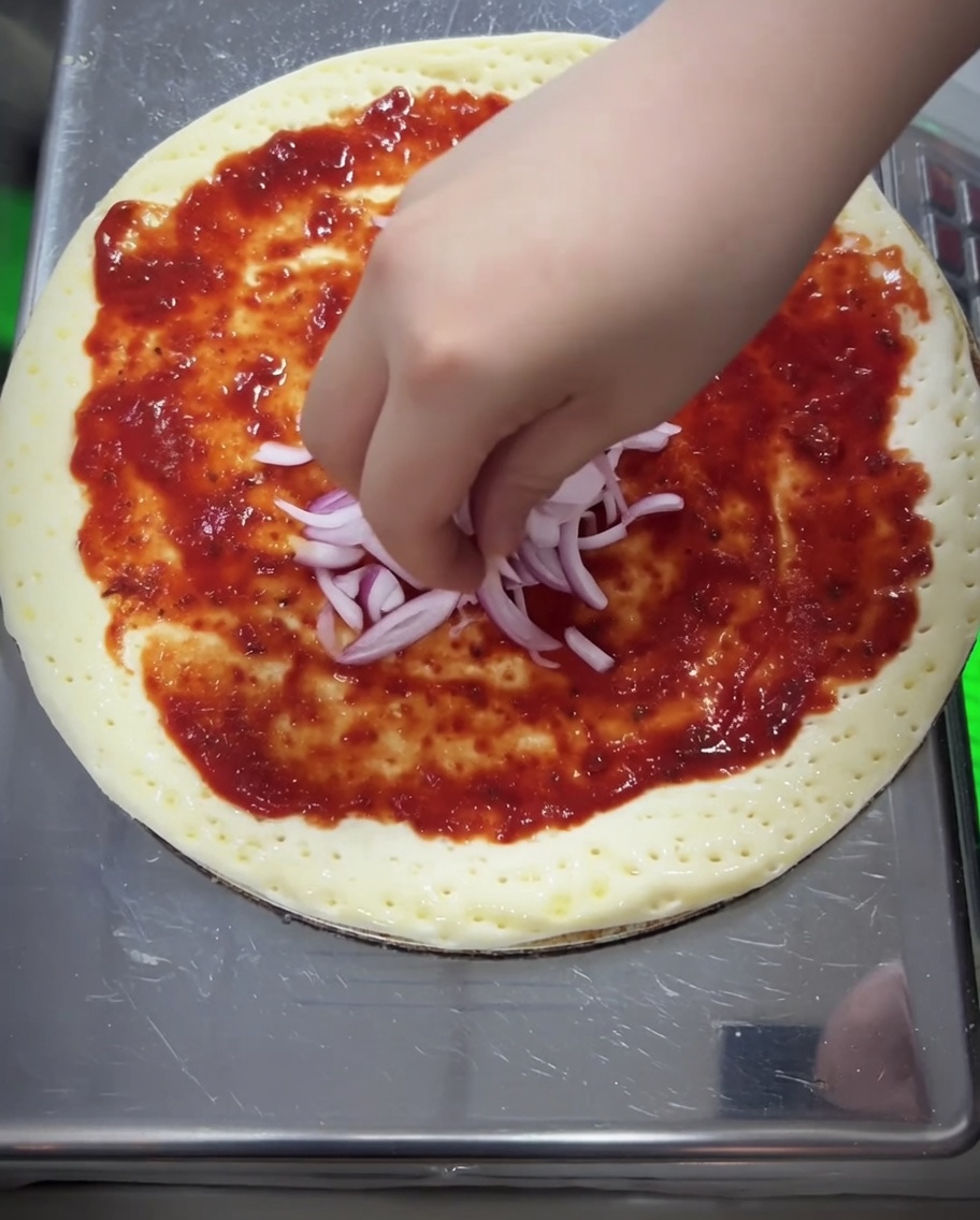 Trozos de cebolla morada y una capa gruesa de queso mozzarella rallado siendo añadidos a la pizza.