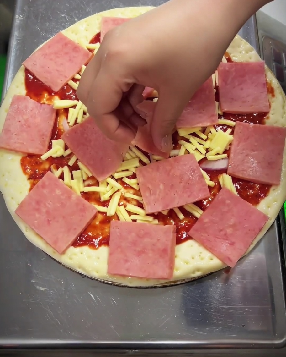 Primer plano de una pizza siendo cubierta con granos de maíz, pimiento verde y champiñones sobre el jamón.