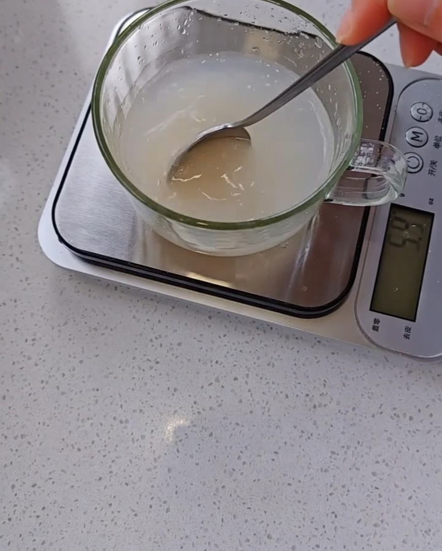Una mano usando una cuchara para remover una mezcla turbia de levadura y agua en una taza medidora de cristal sobre una báscula digital.
