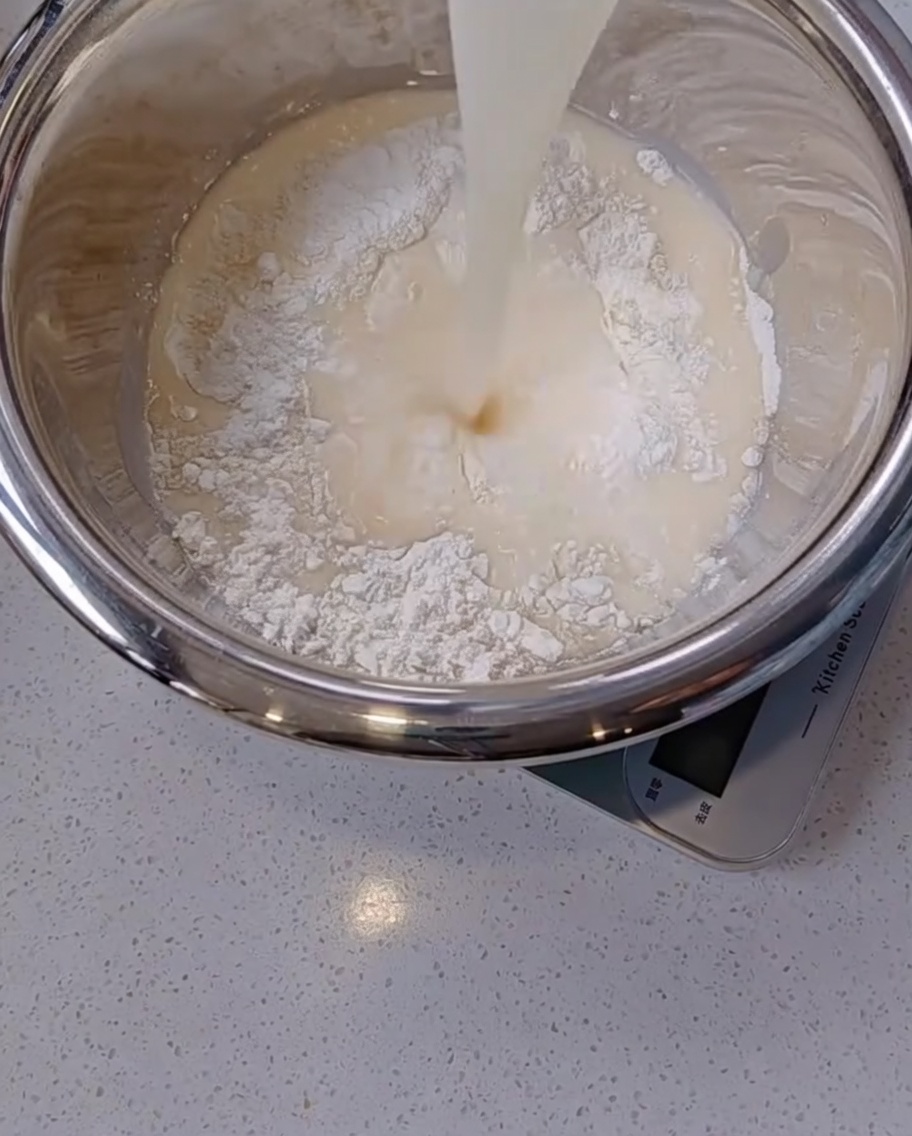 Vertiendo una mezcla lechosa de levadura y agua desde una taza de cristal a un bol de acero inoxidable lleno de harina blanca.