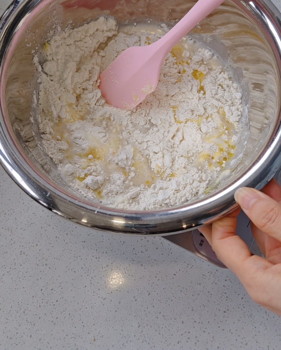 Una mano usando una espátula de silicona rosa para mezclar una masa húmeda y desordenada con restos visibles de aceite de oliva dentro de un bol de acero.