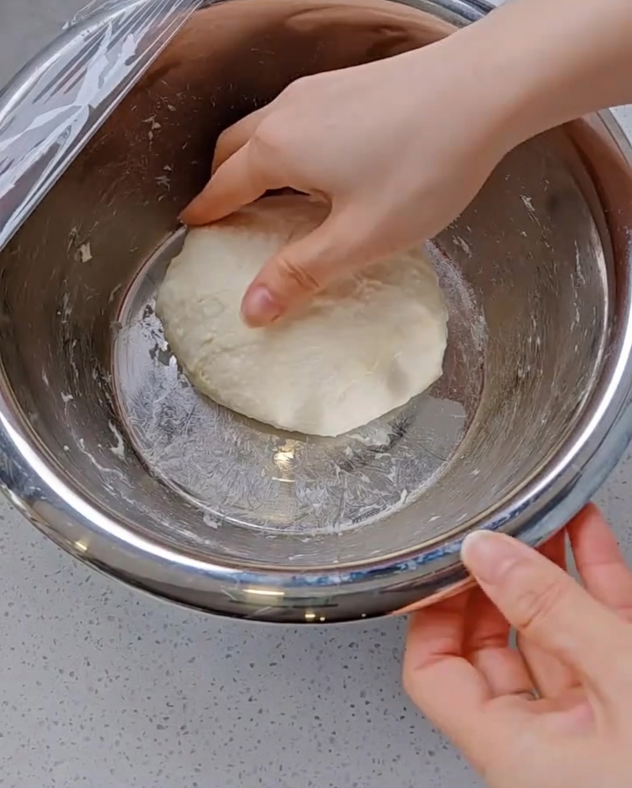 Una mano estirando un trozo de masa de pizza suave y elástica hacia arriba y plegándola hacia el centro dentro de un bol de metal.
