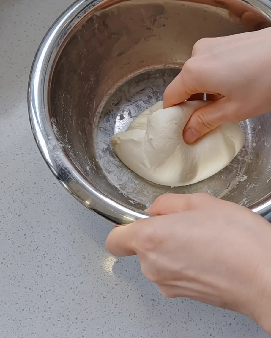Manos metiendo y redondeando suavemente una bola de masa de pizza lisa dentro de un bol de acero inoxidable.