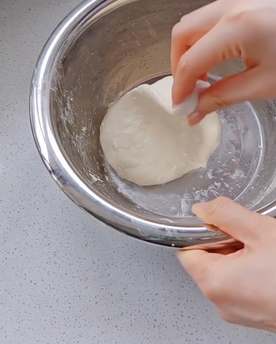 Una mano tirando y estirando un trozo de masa de pizza muy elástica hacia arriba dentro de un bol de metal.
