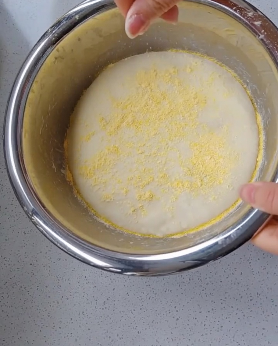 Una mano esparciendo harina de maíz amarilla brillante sobre una bola de masa de pizza hinchada y fermentada dentro de un bol.