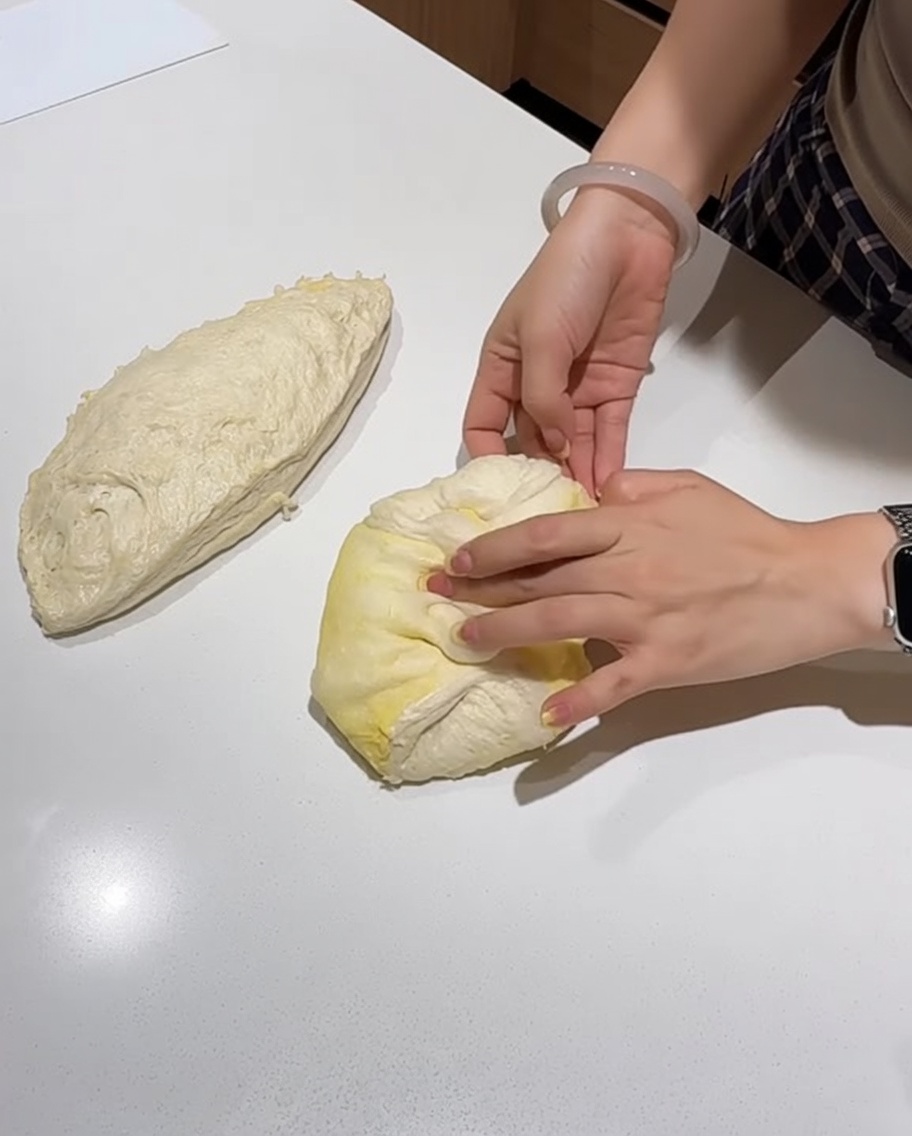 Manos doblando y dando forma a una porción de masa de pizza en un círculo suave sobre una encimera blanca.