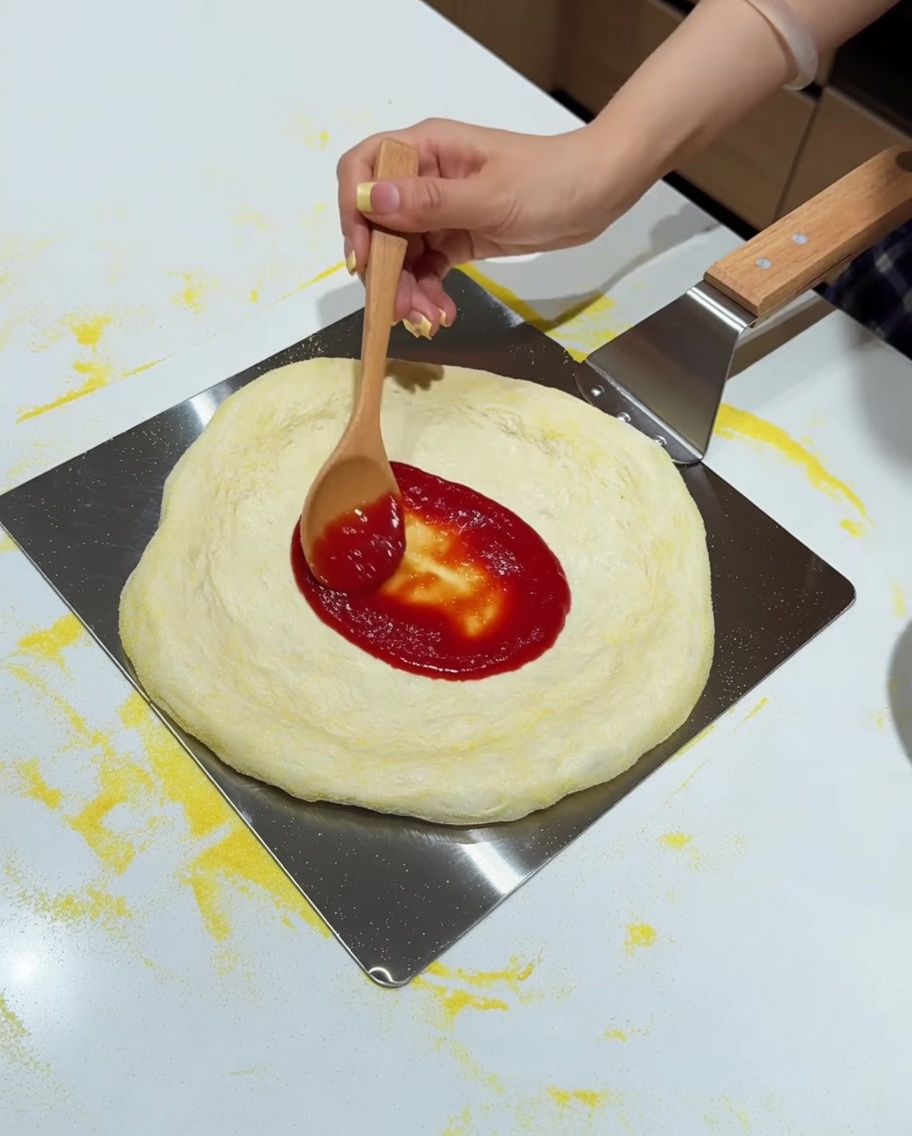 Una cuchara de madera extendiendo salsa de tomate roja sobre una masa de pizza estirada en una pala de metal.