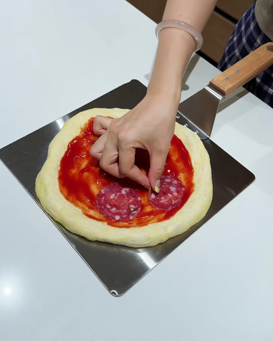 Una mano colocando rodajas de salami sobre una base de pizza con salsa en una pala de metal.