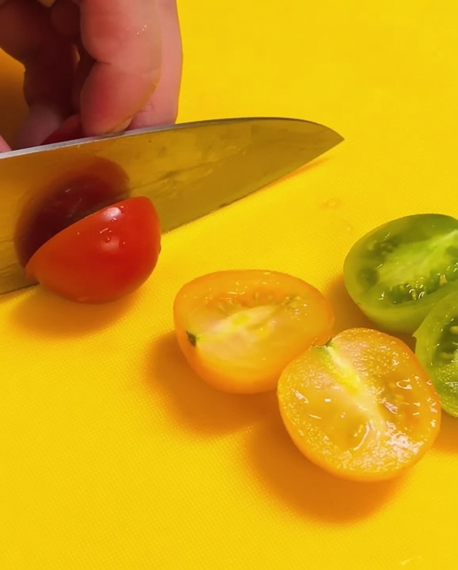 Un cuchillo cortando tomates cherry rojos y verdes por la mitad sobre una tabla de cortar de color amarillo brillante.