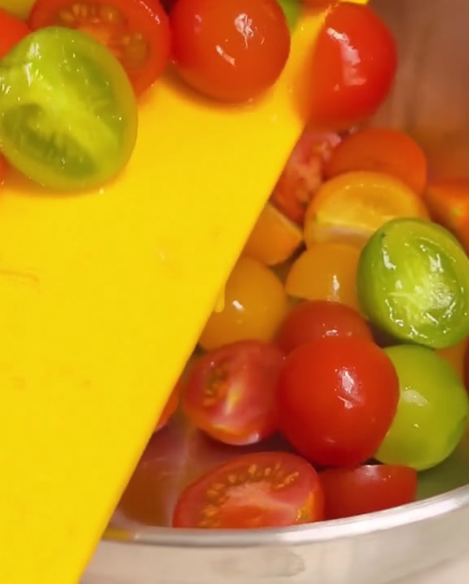 Tomates cherry cortados por la mitad siendo empujados desde una tabla de cortar amarilla hacia un bol de acero inoxidable.
