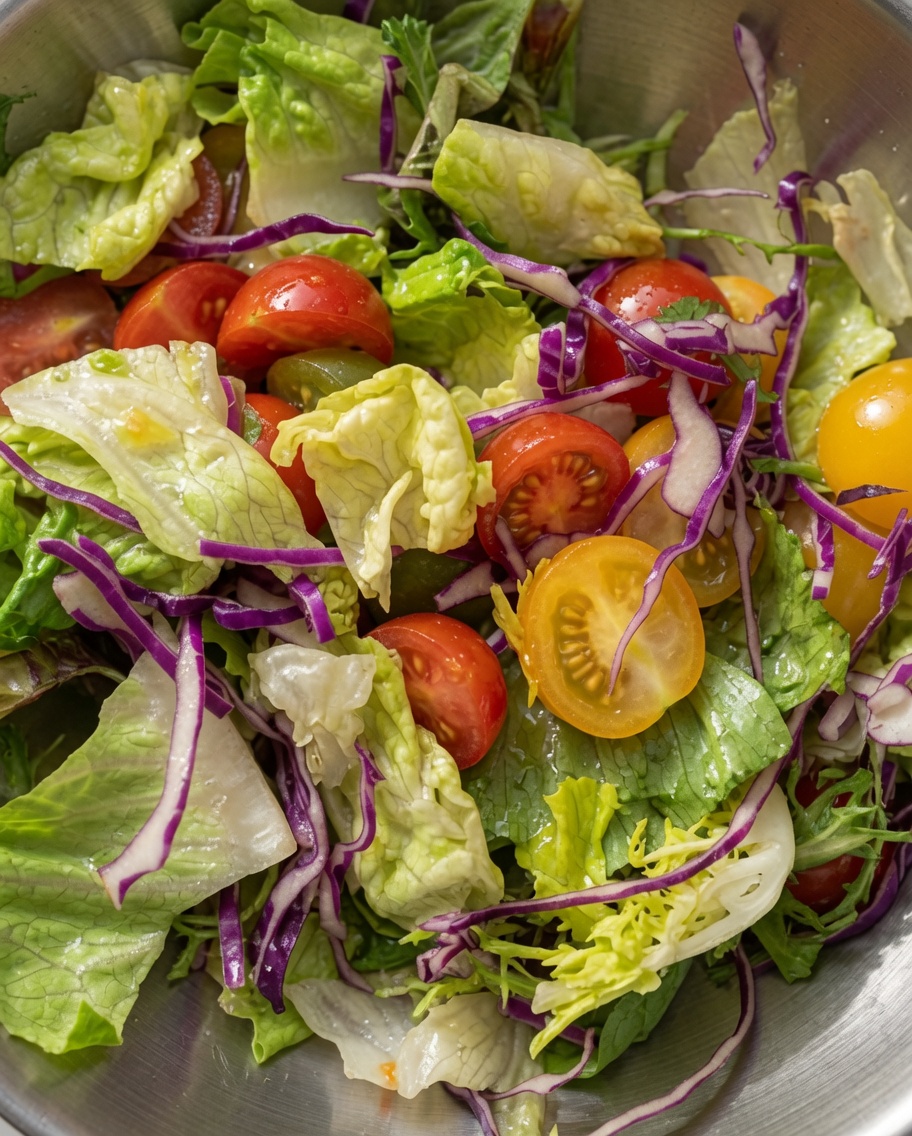 Una vibrante ensalada mixta de lechuga verde, col lombarda y tomates cherry por la mitad, mezclada en un bol de metal.