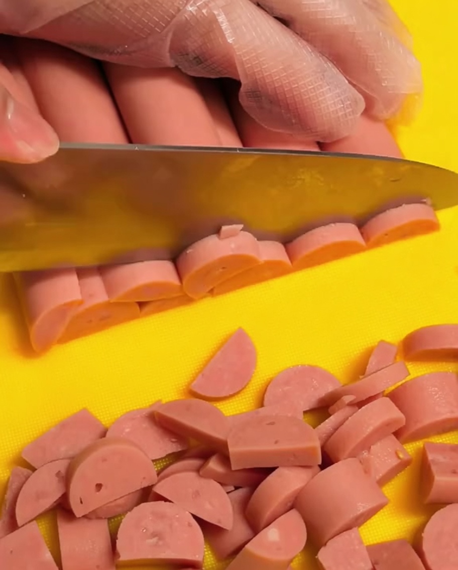 Una mano con un guante transparente cortando salchichas de jamón rosadas en pequeños trozos en forma de media luna sobre una tabla de cortar amarilla.
