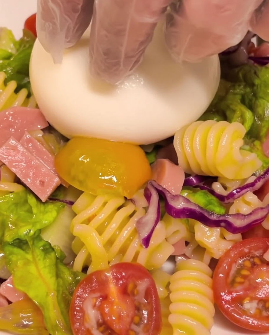 Una bola de queso burrata fresca colocándose en el centro de una ración de ensalada de pasta fría.