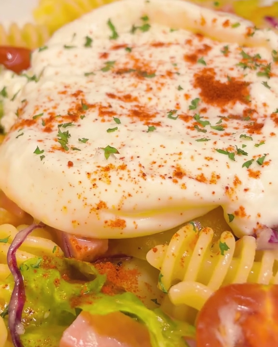 Toma de primer plano del plato de pasta fría terminado con un centro cremoso de burrata, adornado con perejil y pimiento rojo, y un hilo dorado final de aceite de oliva.