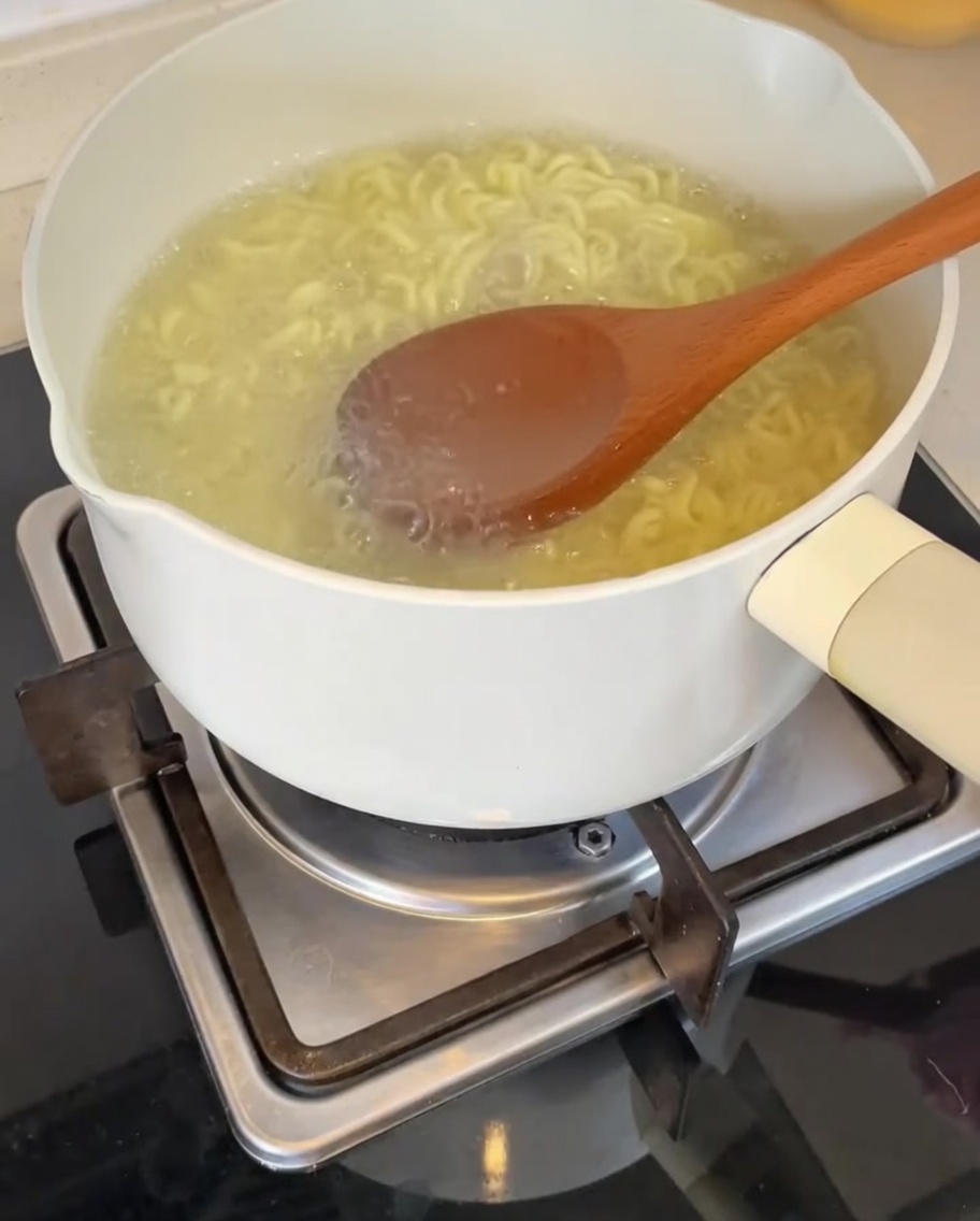 Un cucharón de madera vertiendo agua caliente y turbia de la cocción de los fideos en el tazón que contiene la base de salsa preparada.