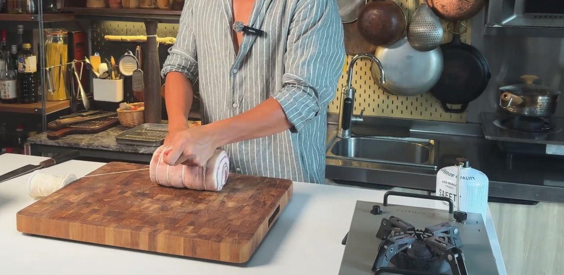 Panceta de cerdo cruda enrollada atada con hilo de cocina sobre una tabla de cortar de madera.