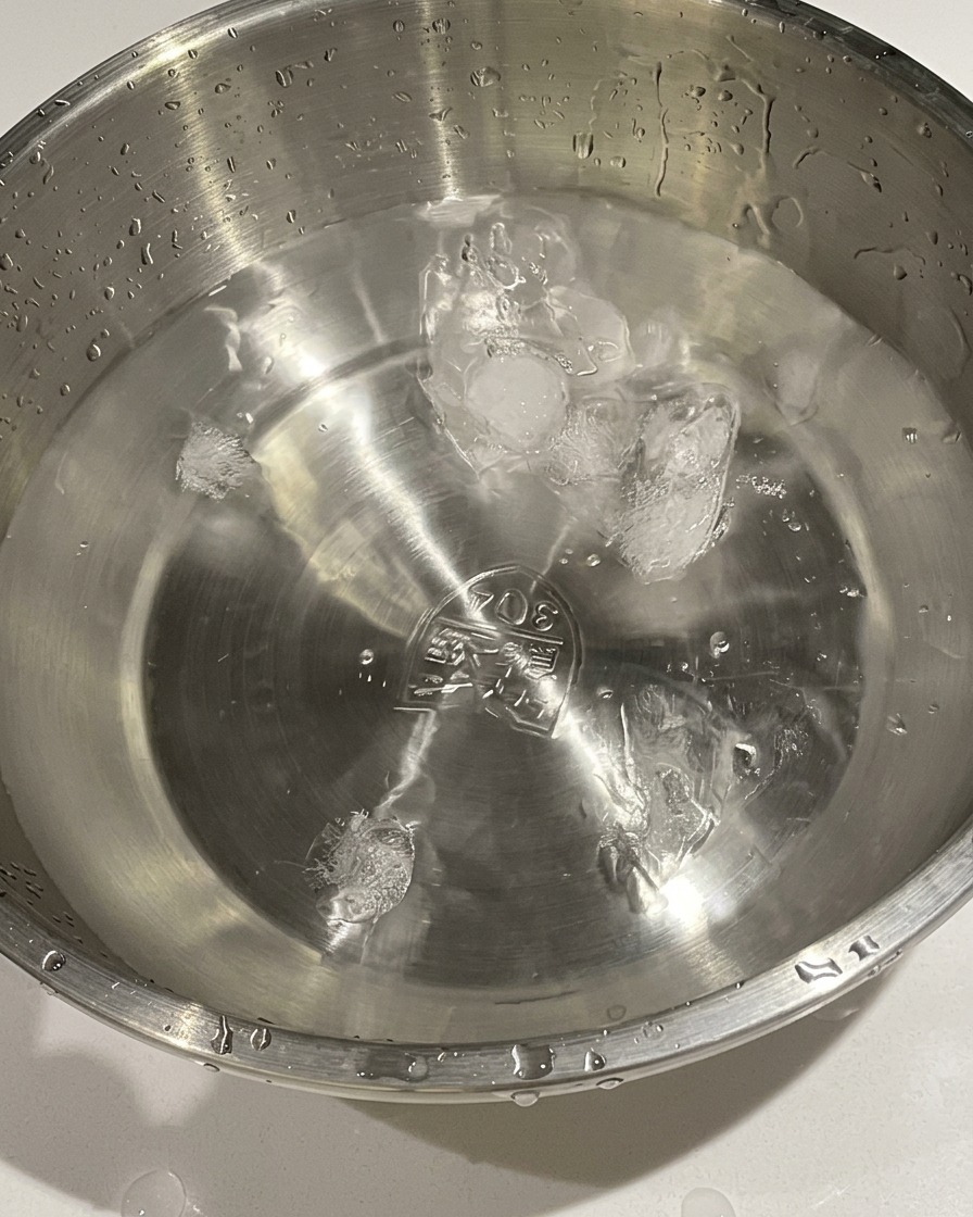 Huevos cocidos siendo transferidos de una olla a un bol de metal con agua fría y cubitos de hielo.