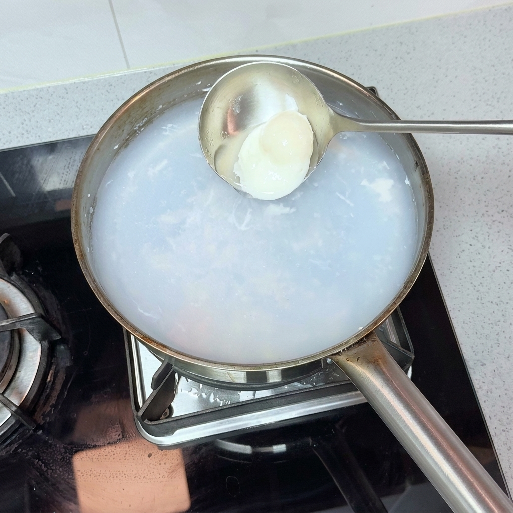Un cucharón de metal recogiendo suavemente un huevo poché perfecto de una olla de agua caliente.