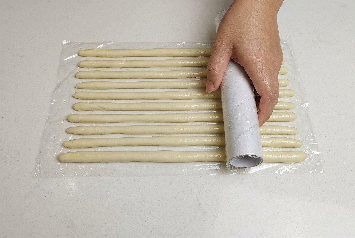 Una mano cubriendo las cuerdas de masa delgadas con papel film sobre una superficie blanca.