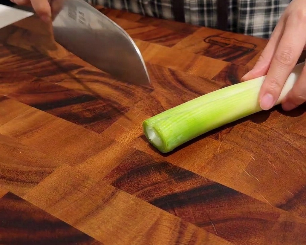 Manos cortando cebollino en rodajas finas sobre una tabla de madera.