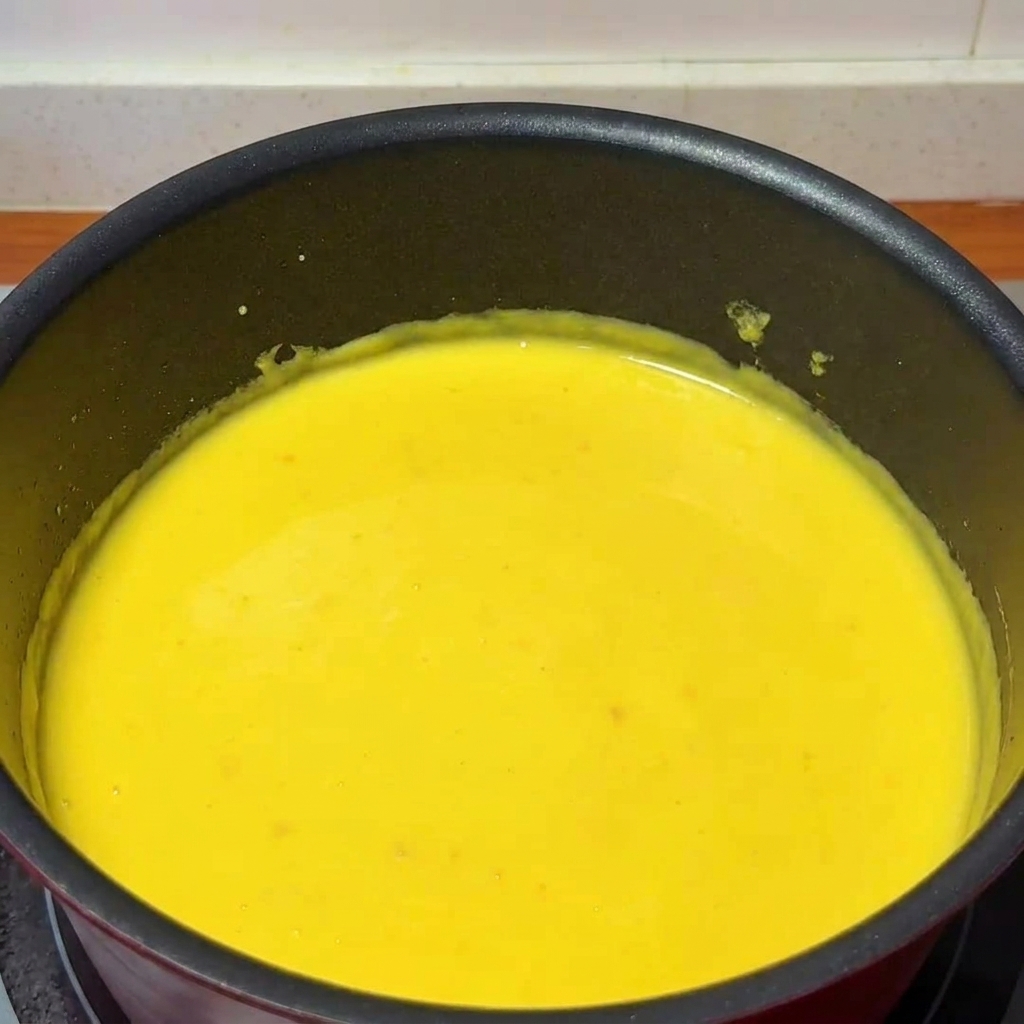 Un cucharón vertiendo zumo de yuzu en un cazo con salsa de queso mientras se remueve con una cuchara de madera.