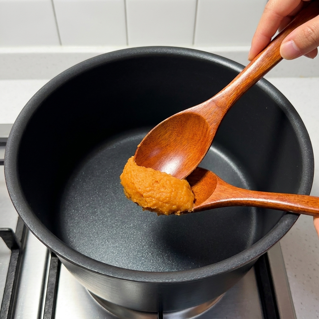 Una cuchara removiendo pasta de miso, mirin, azúcar, caldo de cerdo y zumo de yuzu en un cazo.