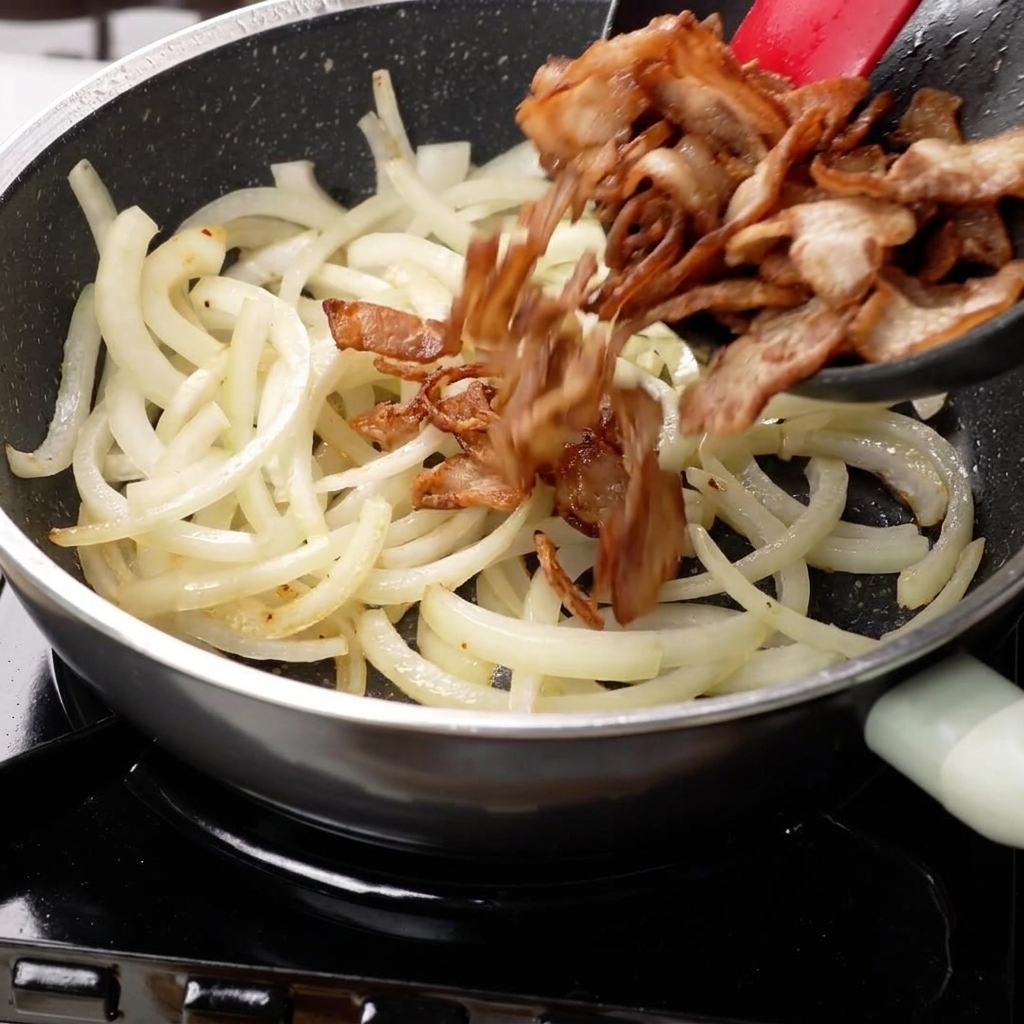 Cebollas salteadas y panceta dorada mezclándose en la sartén.